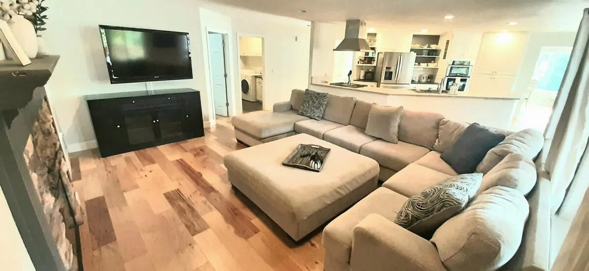 This image features a spacious living area with a large sectional sofa facing a TV and a central coffee table, set in a bright room. The layout is open and clearly designed for comfort, socializing, and entertainment, with ample seating to accommodate a group.