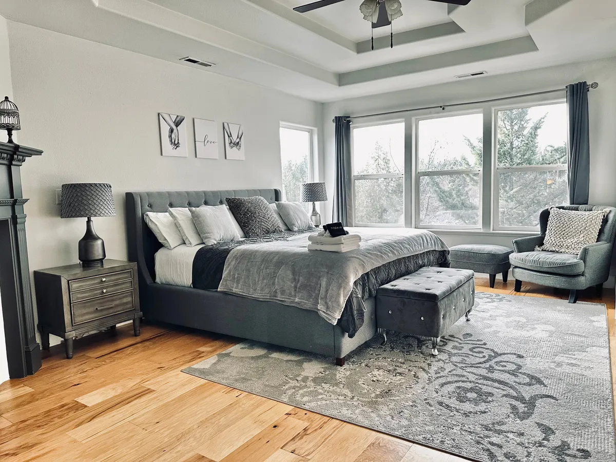 A tranquil bedroom boasts a king-sized bed, large windows with morning light, an armchair perfect for relaxing, a decorative rug, and a nightstand. The room invites moments of quiet reflection or intimate conversation.