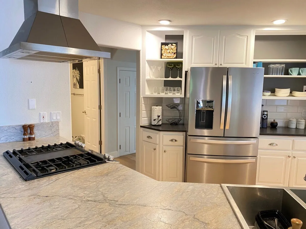 A modern kitchen featuring a stainless steel refrigerator, gas cooktop, spacious island, and open shelving displaying dishware. The kitchen exudes warmth and functionality, offering plenty of space for meal prep and group cooking.