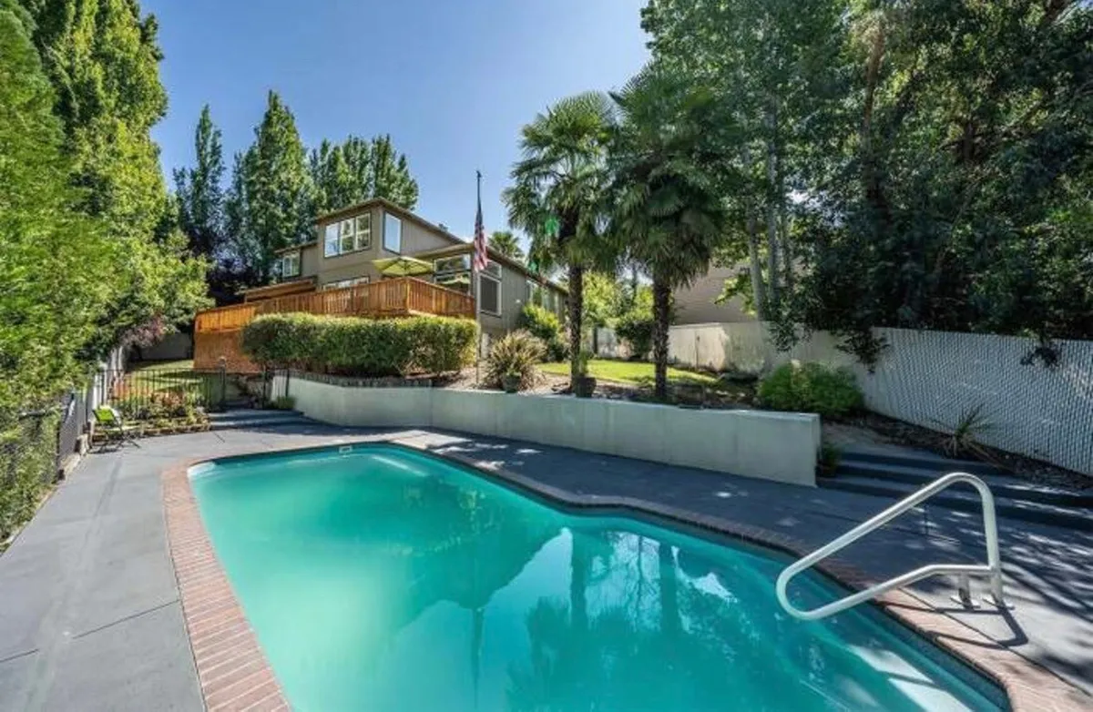 An inviting exterior shot of the home, featuring a sparkling swimming pool surrounded by lush greenery. The house has a balcony overlooking the backyard, offering plenty of space for outdoor gatherings and relaxation. The scene is awash in warm sunset light, suggesting a perfect spot for unwinding after a day of wine tasting or outdoor adventures.