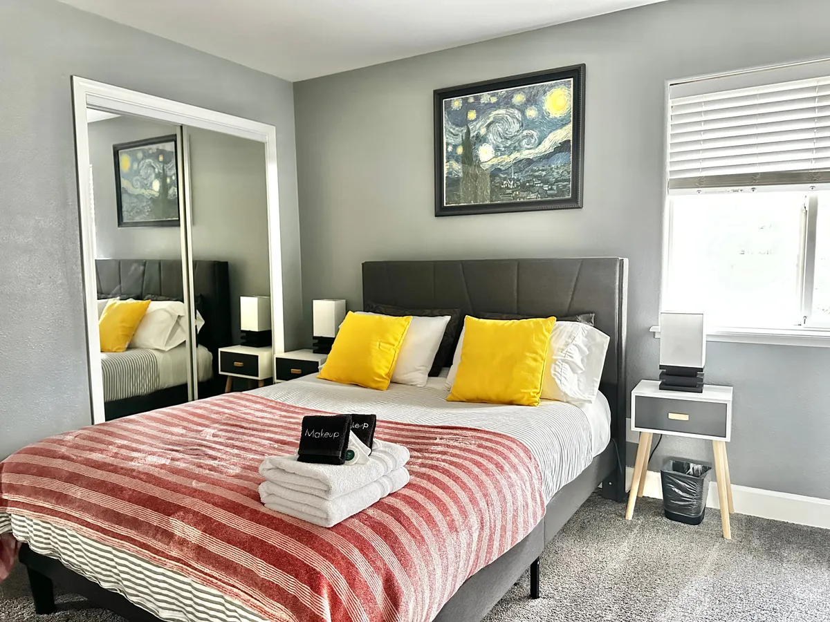 This bedroom scene shows a bed with cheerful yellow pillows, a 'Starry Night' painting above the headboard, a striped blanket, and a nightstand with a lamp. The setting is comfortable and artistic, with a sense of personal style and a cozy place for reading or resting.