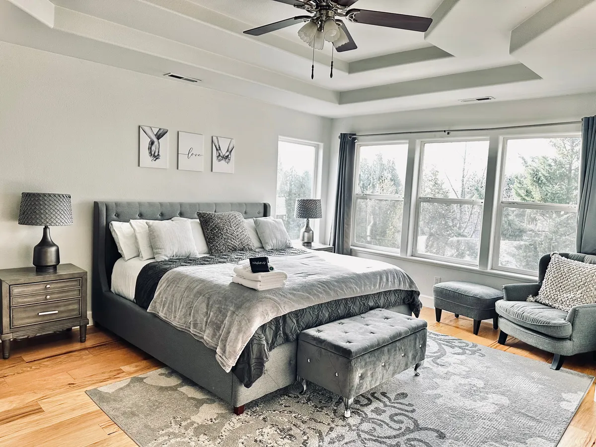 This image features a king-sized bed with plush bedding, large windows bathing the room in natural light, a stylish armchair for reading or relaxing, and decorative artwork on the wall. The overall mood is serene and comfortable, ideal for unwinding after a long day.