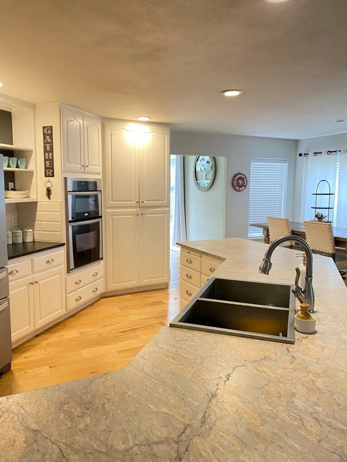 A spacious, light-filled kitchen with a large island, modern sink, and built-in oven. The prominent 'gather' sign over the dining area invites group cooking and meals. The scene captures both the kitchen workspace and the adjacent dining area, emphasizing connection and shared experiences.