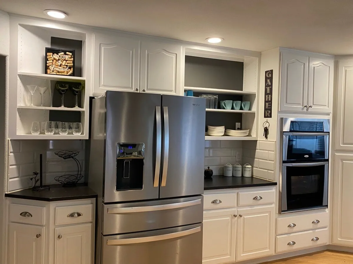 A welcoming kitchen with a black countertop, stainless steel refrigerator, open shelving with glassware, and a decorative 'GATHER' sign. The space feels curated for hosting, blending practical amenities with social touches.